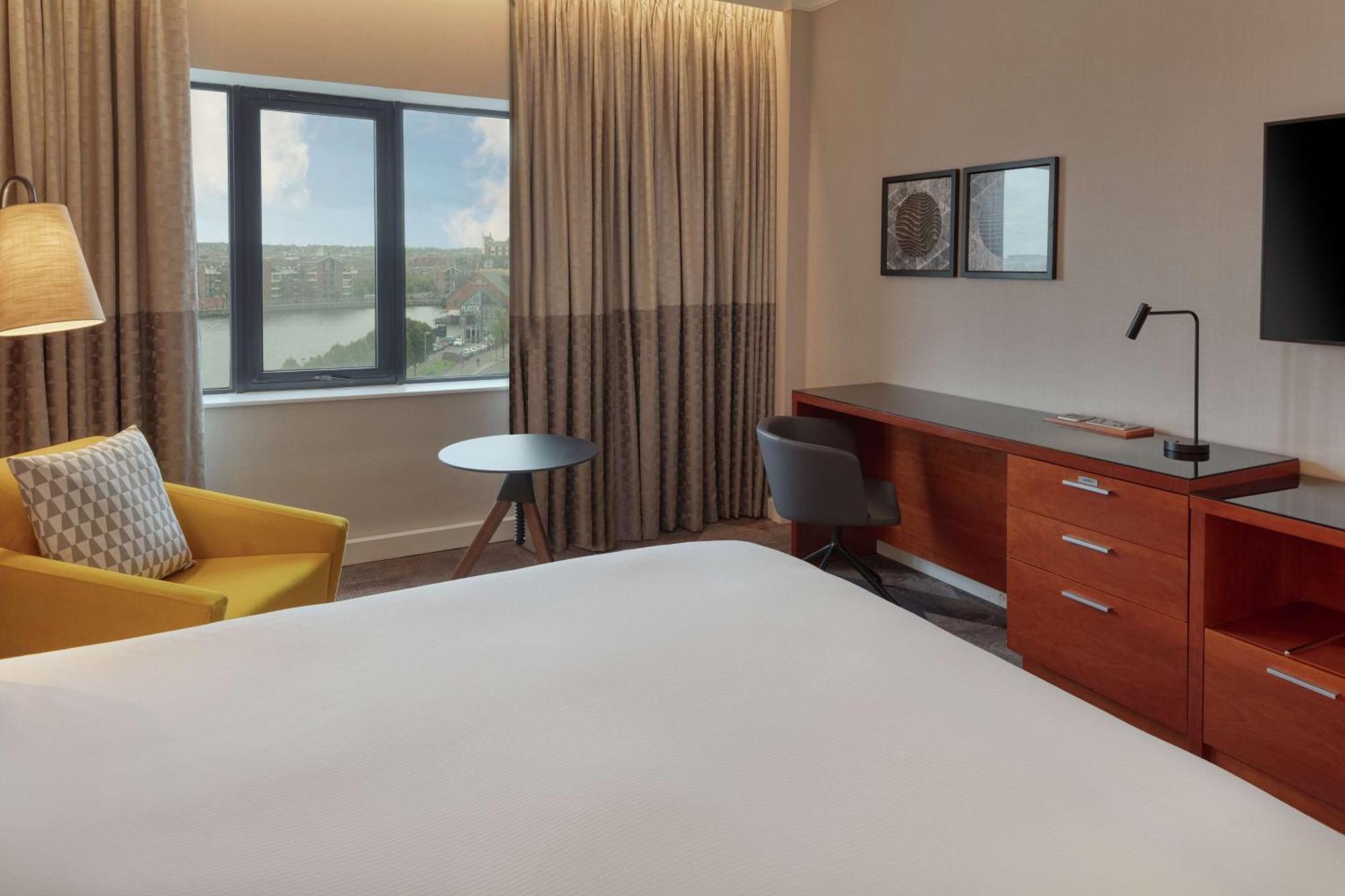 Hilton Belfast Hotel Exterior photo The photo shows a modern hotel room. It features a large bed with a plain white bedspread, and there's a stylish yellow armchair next to it. A small round table is placed nearby, along with a gray chair. The room has large windows covered with light 