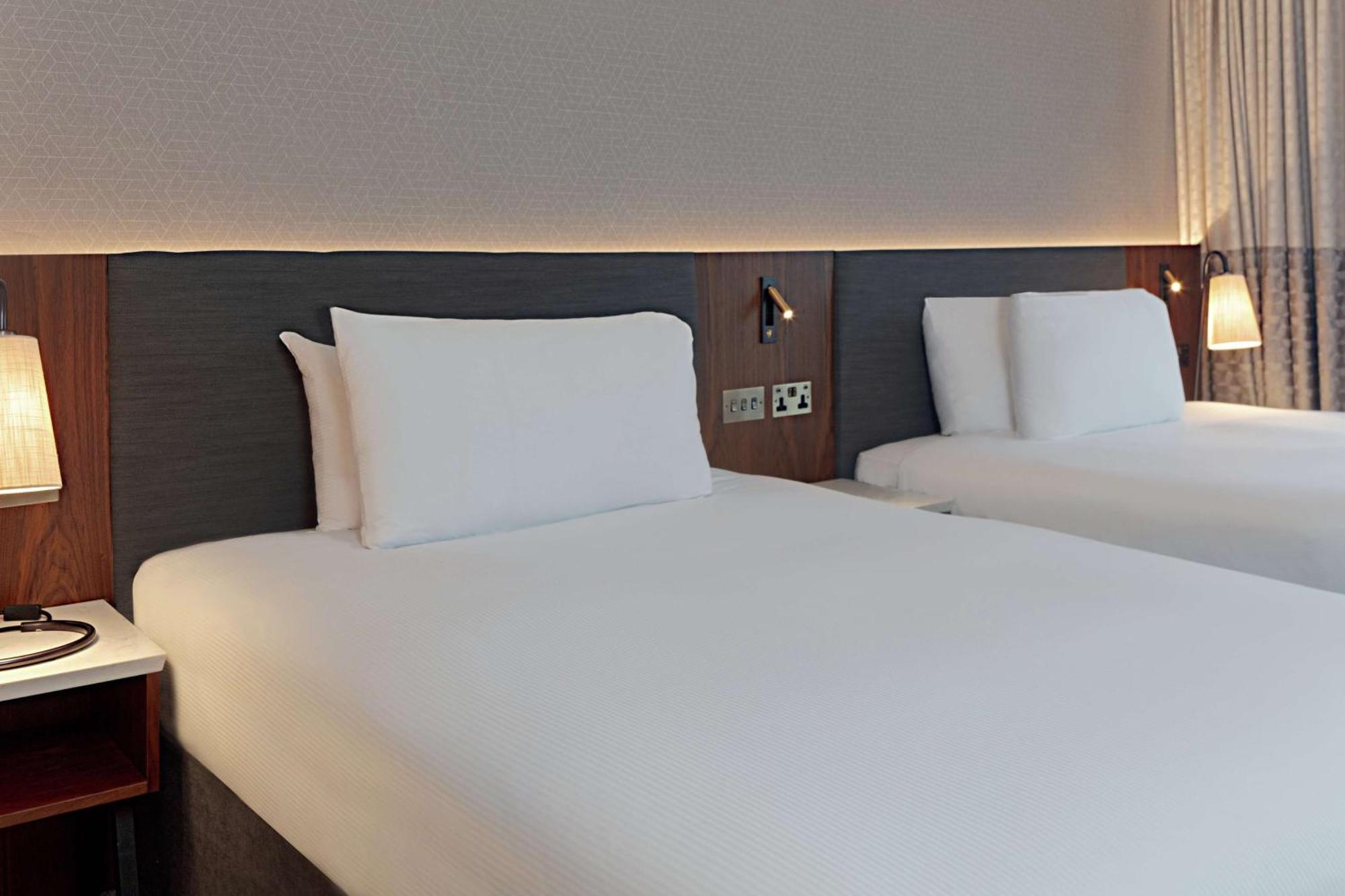 Hilton Belfast Hotel Exterior photo The photo shows a modern hotel room with two neatly made beds. Each bed features white linens and pillows, with a simple yet stylish headboard in a dark wood finish. The room has a soft, ambient light illuminating the area, creating a welcoming atmos
