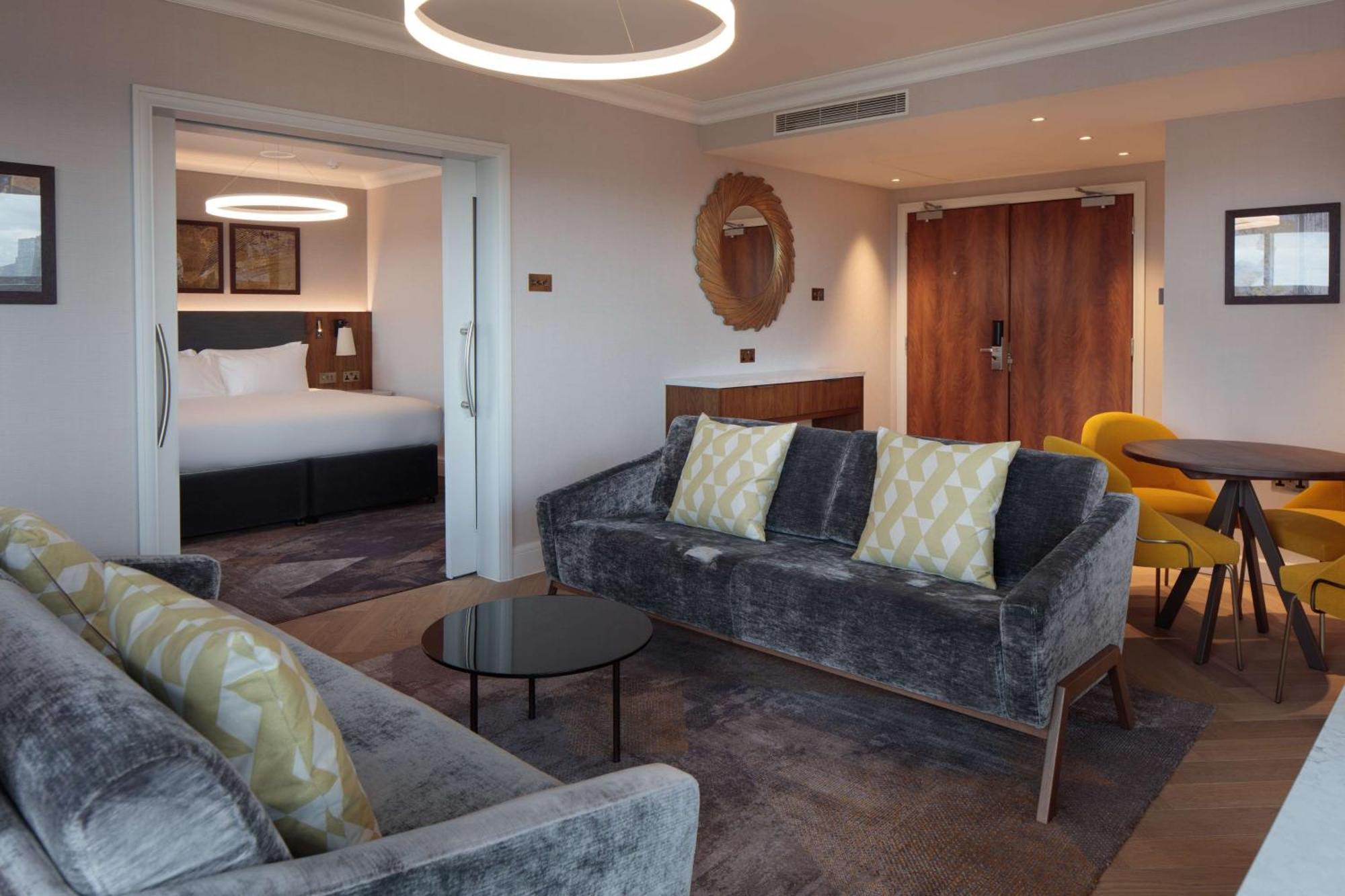 Hilton Belfast Hotel Exterior photo The photo shows a modern and stylish hotel suite. In the foreground, there is a comfortable gray sofa with decorative pillows and a small black coffee table. Adjacent to the sofa is a dining area with a round table and yellow chairs. In the backgroun