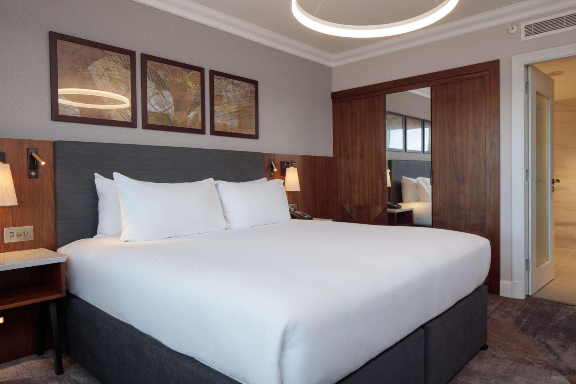 Hilton Belfast Hotel Exterior photo The photo shows a modern hotel room with a clean and contemporary design. The focal point is a large bed with crisp white bedding and two fluffy pillows. The headboard is gray, and next to the bed, there are stylish bedside lamps. Above the bed, ther