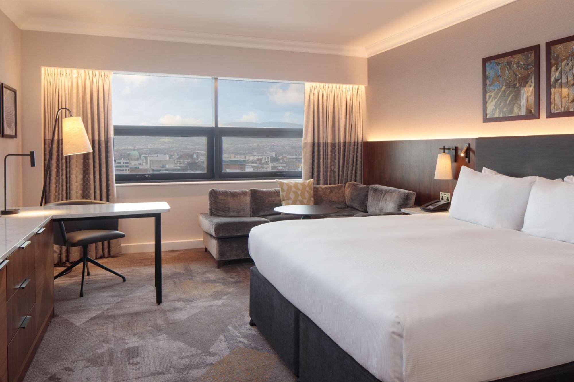 Hilton Belfast Hotel Exterior photo The photo shows a modern hotel room featuring a large bed with white linens positioned against a wall. On one side, there is a small seating area with a couch and a round table. A desk and chair are also present, suggesting a workspace. The room has 