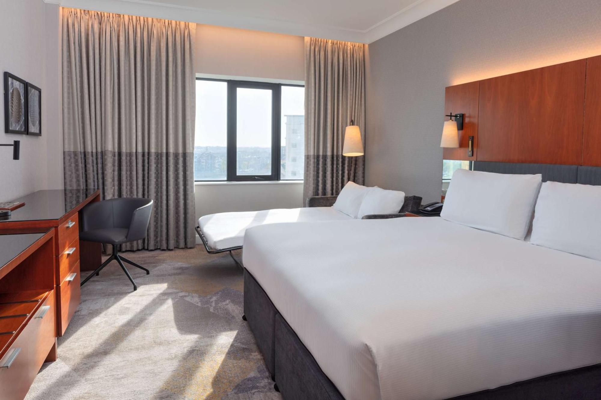 Hilton Belfast Hotel Exterior photo The photo shows a modern hotel room. It features two beds with white linens and a simple, elegant design. There is a large window allowing natural light to enter, covered by light-colored curtains. A comfortable chair and a small desk are placed near