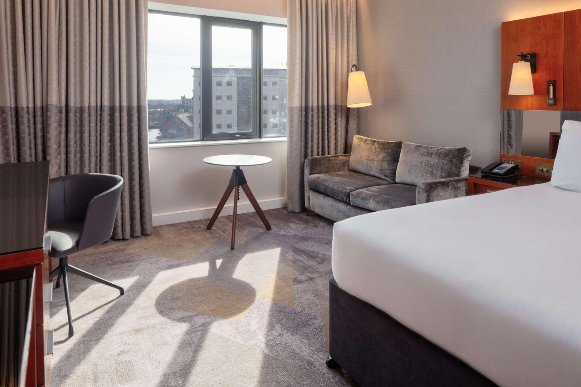 Hilton Belfast Hotel Exterior photo The photo shows a hotel room featuring modern furnishings. There is a large bed with white bedding positioned against one wall. Near the window, which has light-colored curtains, there is a small round table with a tripod-style base. A sleek gray sof