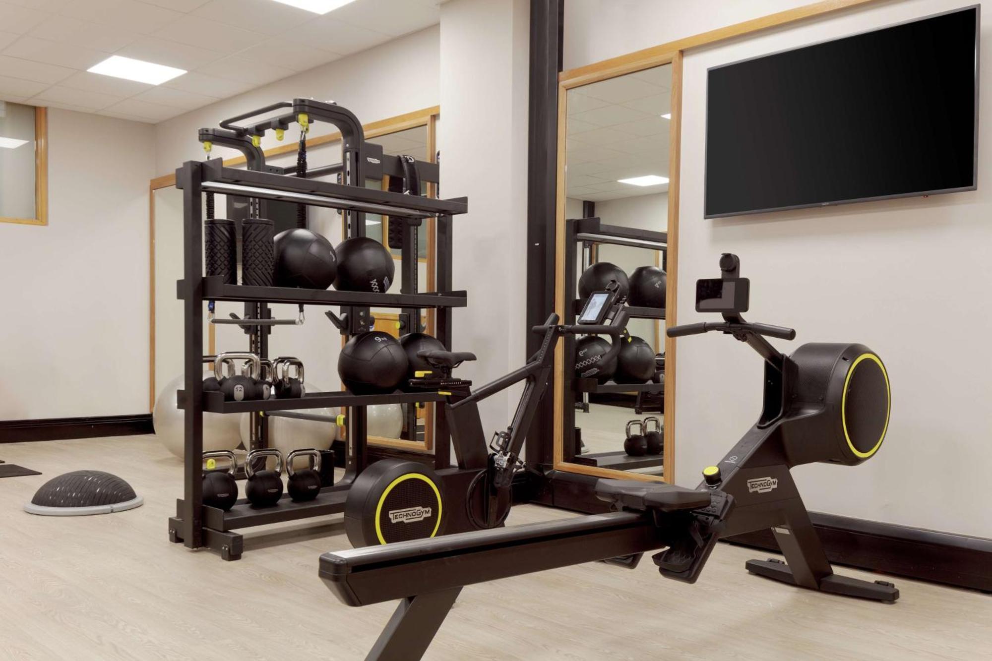 Hilton Belfast Hotel Exterior photo The photo shows a fitness area featuring various exercise equipment. On the left side, there is a rack that holds multiple black kettlebells and a few other fitness accessories. In the center, there is a rowing machine designed for cardio workouts. T