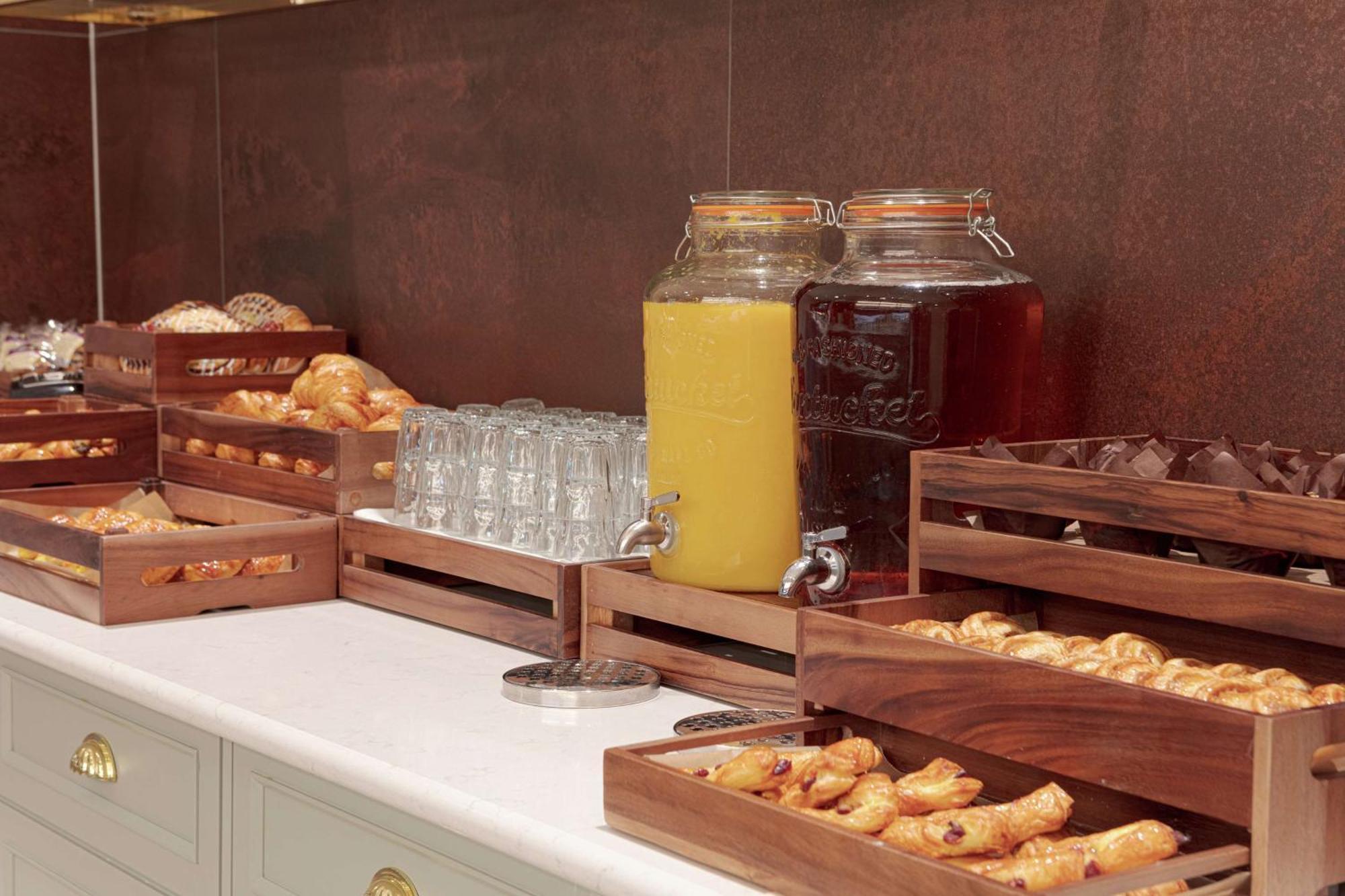 Hilton Belfast Hotel Exterior photo The photo shows a buffet-style setup featuring various food and beverage items. There are several wooden trays filled with pastries, including croissants and other baked goods. In the background, there are two large glass jars, one filled with yellow