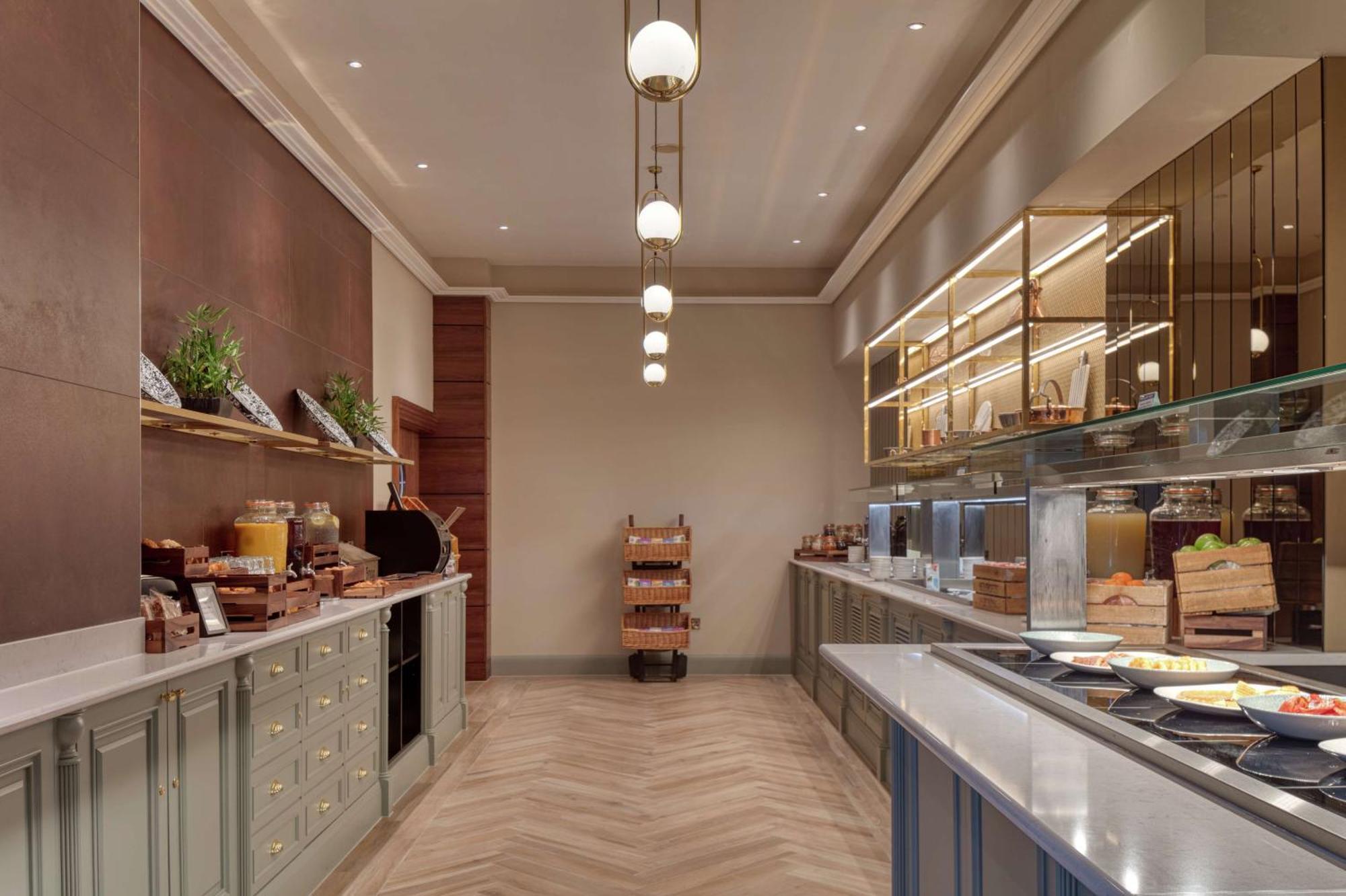 Hilton Belfast Hotel Exterior photo The photo shows an elegantly designed dining area, likely a buffet or breakfast setup. On one side, there are wooden shelves displaying various food items and plants, contributing to a warm atmosphere. Along the opposite side, there's a glass display