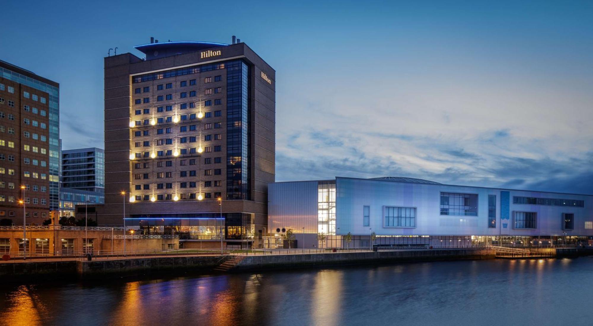 Hilton Belfast Hotel Exterior photo The image shows a Hilton hotel building along a waterway, illuminated during twilight. The hotel features a modern architectural design with multiple stories and large windows. To the right of the hotel, there is another structure that appears to be 